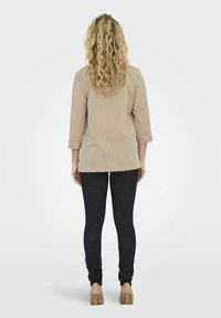 Beige, textuur top met driekwart mouwen, gecombineerd met donkere skinny jeans en lichtgekleurde hakken. De achterkant toont de lengte en pasvorm.