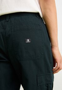 Gros plan d'une personne portant un pantalon Converse noir avec une poche arrière et un logo visible, la main reposant sur le côté.
