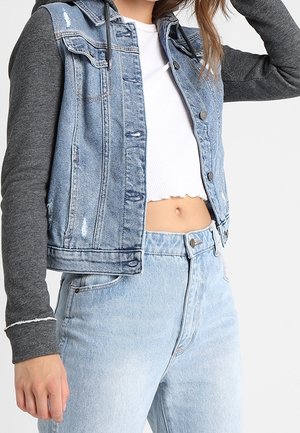 Mujer con jeans de mezclilla claros, top blanco corto de canalé y chaqueta de mezclilla con mangas de sudadera gris oscuro, recortada a la altura de la cintura.