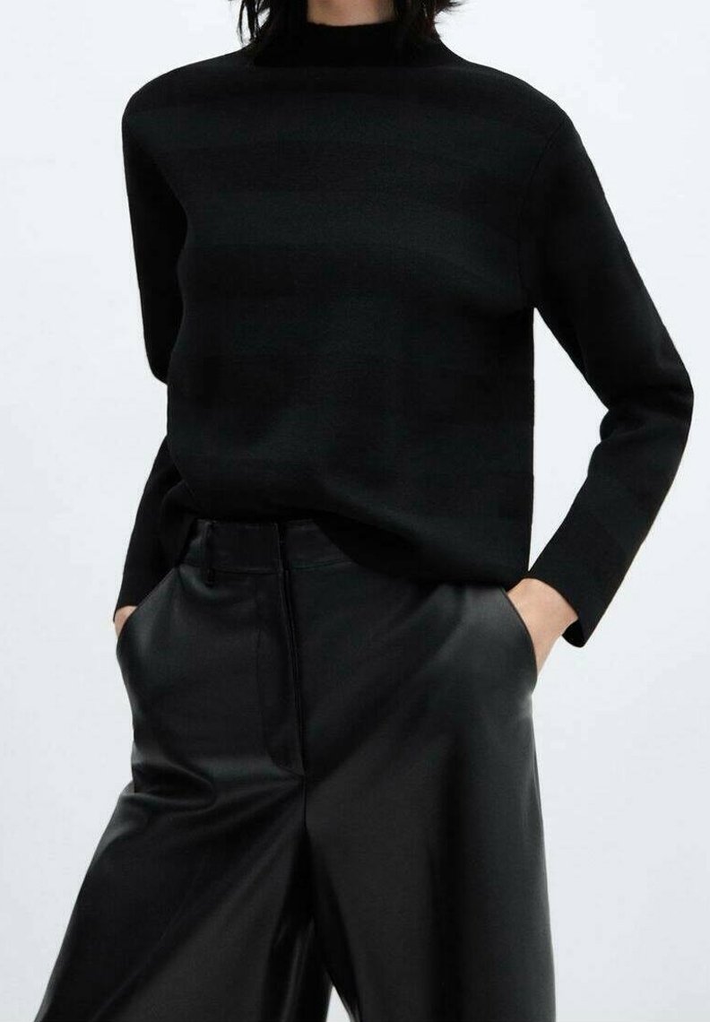 Personne portant un pull noir à manches longues et un pantalon large en cuir noir taille haute, les mains dans les poches.