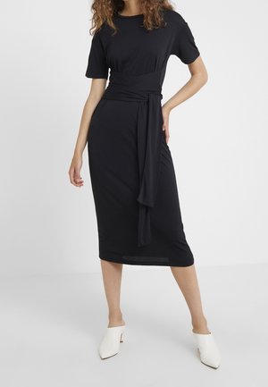 Vrouw draagt een aansluitende zwarte midi-jurk met korte mouwen en een ceintuur gestrikt in de taille, gecombineerd met witte hak-mules, staand tegen een effen achtergrond.