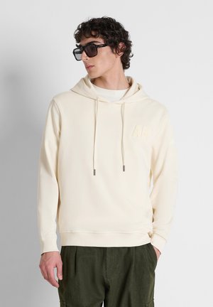 Man draagt een crème hoodie met koordjes en donkergroene broek, poseert met handen in de zakken, kijkt opzij terwijl hij een zwarte zonnebril draagt.