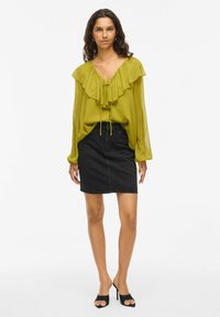 Limettengrüne, gerüschte Bluse mit Kordelzug am Hals, kombiniert mit einem schwarzen Jeansrock. Das Model trägt schwarze offene High Heels.