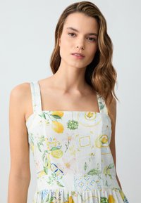 Wit bloemenjurk met gele citroen- en groene leliepatronen, vierkante halslijn, brede bandjes en een geplooide rok-textuur.