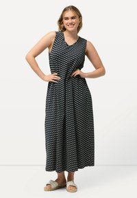 Černé maxi šaty bez rukávů s bílým geometrickým vzorem, výstřih do V, nabíraný pas a volná sukně. Nosené s lehkými sandály.
