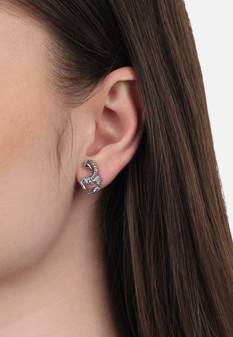 Arete en forma de escorpión plateado con textura y diseño detallado, sujeto al lóbulo, llevado en una vista de cerca junto a cabello largo y liso.