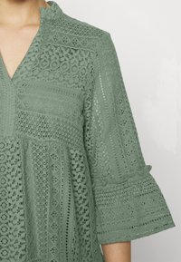 Bluză verde din dantelă cu decolteu în V, manșete evazate până la trei sferturi, modele intricate și un finisaj texturat.