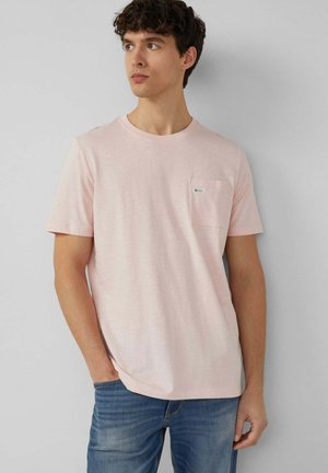 Młody mężczyzna o kręconych włosach, ubrany w jasnoróżowy t-shirt z krótkim rękawem i kieszenią na piersi oraz niebieskie dżinsy, stojący przy jasnoszarej ścianie.