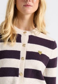 Stripet cardigan i lys creme og mørk lilla, med guldknapper og et lille gyldent hjertepin på brystet. Uldblanding.