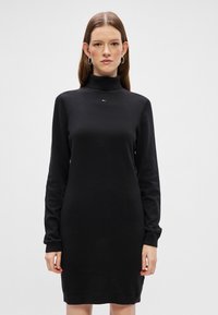 Vestido de pulôver preto com gola alta, mangas longas e bainha canelada. Apresenta um pequeno logotipo centrado no peito. Textura lisa.