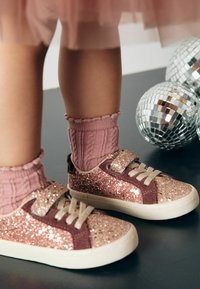 Glitzernde rosa und braune Turnschuhe mit einem Klettverschluss, kombiniert mit gerippten rosa Socken. Der Hintergrund zeigt kleine Discokugeln.