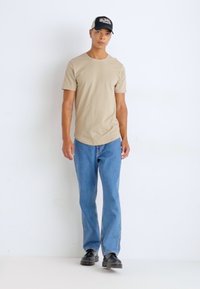 T-shirt beige a maniche corte abbinato a jeans blu chiaro a gamba dritta e scarpe nere, indossato con un cappellino nero con dettagli bianchi.