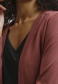 En nära bild av en person som bär en vinröd cardigan över en svart topp, som vilar en hand på hakan, med lockigt hår synligt.