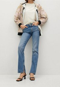 Jasnoróżowa kurtka w wzory z czarnymi akcentami, biała koszulka oraz proste spodnie dżinsowe w średnim odcieniu niebieskiego, zestawione z czarnymi sandałami na obcasie.