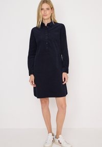 Femme se tenant contre un fond blanc, portant une robe chemise bleu marine à manches longues et des baskets blanches avec des accents rouges.