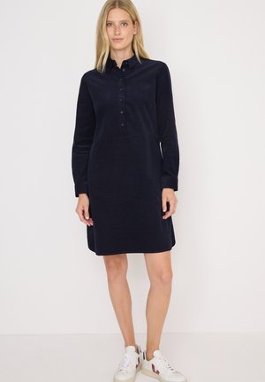 Femme se tenant contre un fond blanc, portant une robe chemise bleu marine à manches longues et des baskets blanches avec des accents rouges.