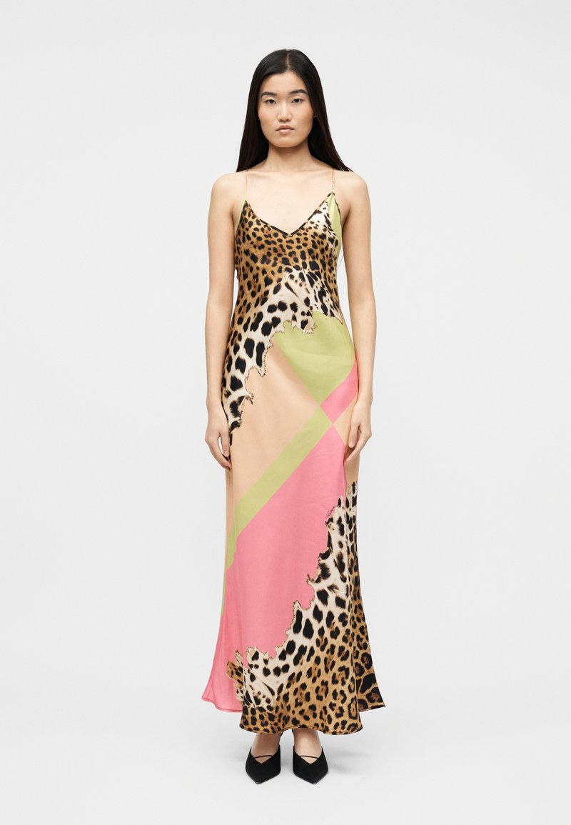 Vrouw staand, gekleed in een lange slipjurk met luipaardprint gecombineerd met roze en groene geometrische patronen, gecombineerd met zwarte puntige schoenen.