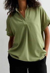 Short-sleeve, collared shirt in light green with a textured, grid pattern. Loose fit with a subtle pleat at the front.