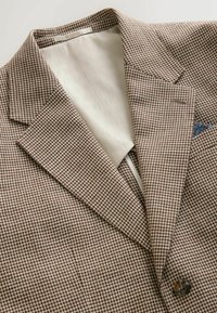 Svetlohnedé a krémové sako s kockovaným vzorom houndstooth, so zárezovým klopou, jednoplášťové s jedným gombíkom, s krémovou vnútornou podšívkou a modrým vzorovaným vreckovkom v hrudnom vrecku.