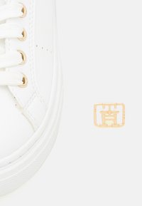 Zapatilla blanca con ojales y cordones dorados, suela de goma texturizada y un logo emblema dorado junto al zapato.