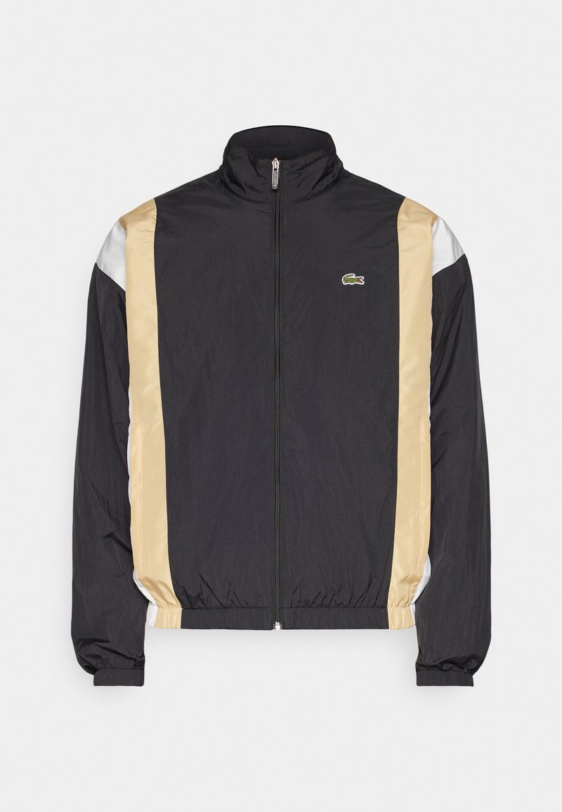Veste coupe-vent noire avec des panneaux latéraux en satin beige, accents argentés, col montant et poignets élastiques. Présente un petit logo sur la poitrine.