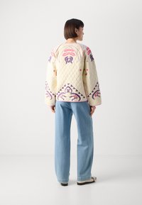 Quiltet kremfarget jakke med rosa og lilla blomsterbroderi, med vide ermer og kant. Kombinert med lyseblå jeans og svarte sko.