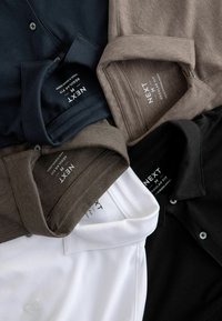Vijf poloshirts in marineblauw, bruin, wit en zwart, met een reguliere pasvorm en knoopkragen. Labels tonen het merk "NEXT".