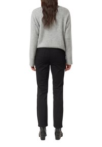 Osoba s dlouhými hnědými vlasy, oblečená v světle šedém svetru, černých kalhotách a černých vysoce podpatkových kotníkových botách, stojící zády.