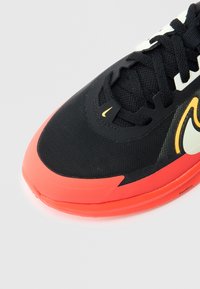 Μαύρο αθλητικό παπούτσι με υφασμάτινο επάνω μέρος με υφή, φωτεινή κοραλί σόλα και διακριτικές κίτρινες λεπτομέρειες. Διαθέτει παραδοσιακά κορδόνια και επένδυση στον γιακά.