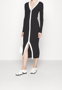 Abito a maglia nero a righe, con maniche lunghe, scollatura a V, chiusura con bottoni bianchi e spacco laterale. Abbinato a sandali piatti bianchi. Adatto per un look casual.