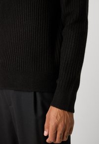 Pulover negru tricotat cu model côtelé, cu mâneci lungi, având un model texturat și un manșon ajustat. Purtați-l cu pantaloni negri.