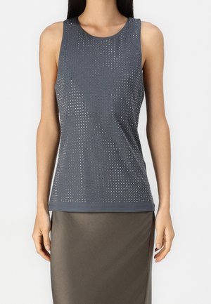 Frau, die ein ärmelloses dunkelgraues Tanktop mit einem Gitter aus kleinen metallischen Nieten trägt, kombiniert mit einem dunkelgrauen Rock.
