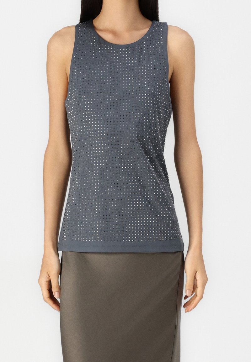 Vrouw draagt een mouwloze donkergrijze tanktop met een raster van kleine metalen studs, gecombineerd met een donkergrijze rok.