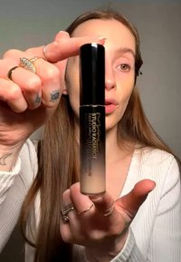 Frau mit langen Haaren hält eine Flasche MAC Studio Radiance flüssigen Concealer und zeigt sie nah an der Kamera vor einem einfarbigen Hintergrund.