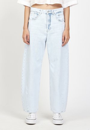 Woman wearing light blue high-waist wide-leg jeans, white cropped top, and white sneakers, standing against a plain background.