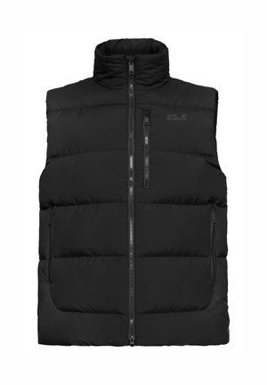 Junger Mann trägt schwarze Steppweste, rotes Langarmshirt, schwarze Hose, schwarze Schuhe und dunkle Sonnenbrille, steht mit den Händen in den Taschen.