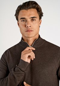 Sweater castanho com fecho zip, com gola e punhos canelados, apresentando uma textura suave e um design justo. Modelo a segurar o fecho.