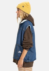 Person som har på seg en gul lue, brun hettegenser, blå ermeløs denimjakke og rutete bukser, står i profil mot hvit bakgrunn.