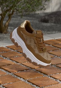 Baskets Guess marron et beige avec semelle blanche épaisse et détails dorés, présentées sur un sol extérieur en carreaux de bois et galets.