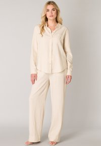 Beige linnen overhemd en bijpassende brede broek. Het overhemd heeft een knoopsluiting aan de voorkant en een borstzak. Zachte, textuurstof.