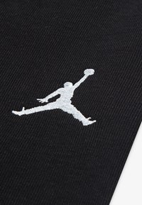 A fekete sportos anyagon egy hangsúlyos, fehér hímzett Jumpman logó látható, amely egy, kosárlabdát dobni készülő alakot ábrázol.