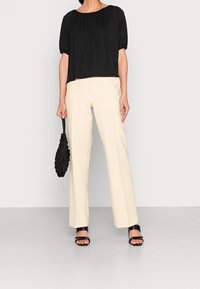 Sort top med puffærmer, parret med cremede buksestil med brede ben. Modellen holder en tekstureret sort taske og har sorte strophæle på. Minimalistisk design.