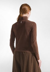 Brun bluse med lange ærmer og høj krave, blød tekstur og et diskret mærke bagpå, sat sammen med en matchende brun nederdel.