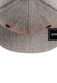 Grå caps med strukturet stoff, buet skygge og en svart etikett med merkevarenavnet "MSTRD." Sirkulært hull for ventilasjon på toppen.