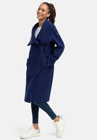 Manteau long bleu marine avec col châle, fermeture éclair à l'avant, poches latérales et texture douce, associé à un jean et des baskets blanches.