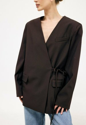 Woman wearing an oversized black wrap blazer with front flap pockets and a side tie, paired with light blue jeans.