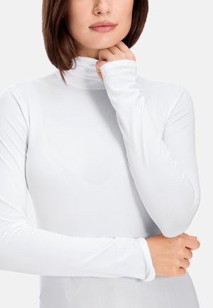 Woman with brown hair wearing a white long-sleeve turtleneck top, resting her chin on one hand and crossing her arms.