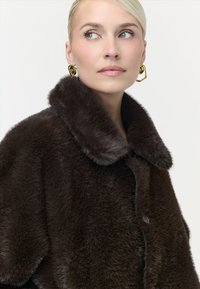 Femme blonde aux yeux bleus portant des boucles d'oreilles en or et un manteau duveteux marron foncé, regardant légèrement vers la droite sur un fond blanc.
