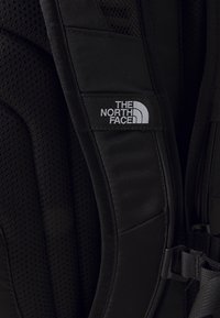 Közelkép egy fekete párnázott hátizsákpántjáról, amelyen a The North Face logója fehér színben a pánt felső részén található.