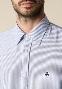 Detailní záběr dolní části obličeje muže a krku, který má na sobě modro-bíle pruhovanou košili s malou vyšívanou nášivkou na hrudi.